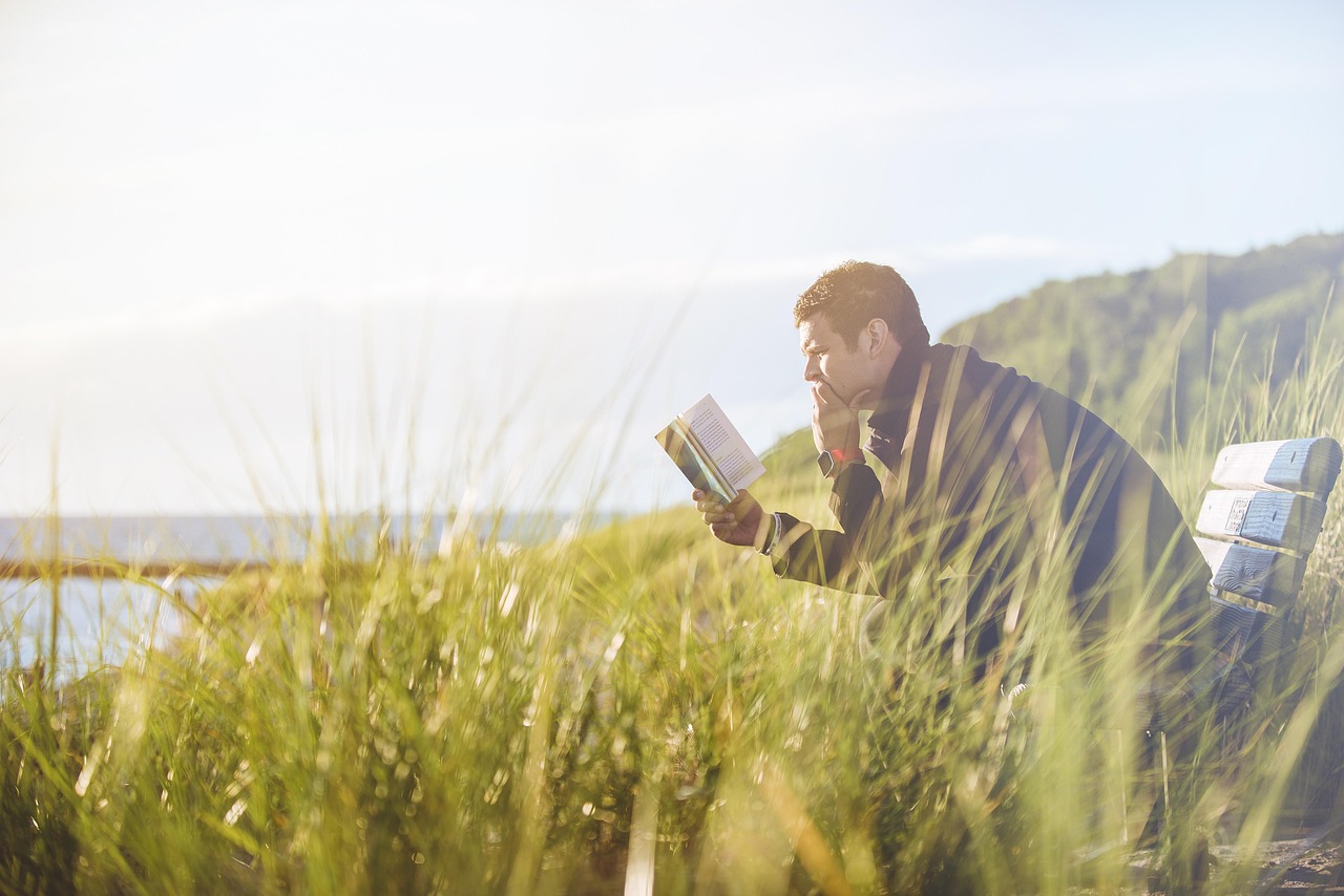 March Winner: Portrait of a man sitting on a bench in tall grass by the water, engrossed in reading a book in bright sunlight.